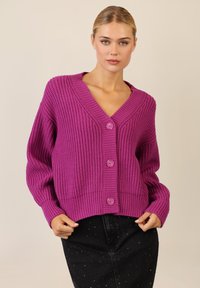 Paarse gebreide cardigan met een diepe V-hals, voorzien van textuurribbeling en drie voorknopjes. Gecombineerd met een donkere rok.