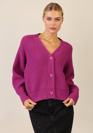 Purple knitted cardigan with a deep V-neck, featuring textured ribbing and three front buttons. Paired with a dark skirt.