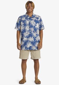 Camisa hawaiana de manga corta azul con patrones de hojas blancas, combinada con pantalones cortos de pana beige y sandalias de tiras marrones.