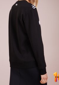 Sudadera negra con cuello redondo y mangas largas. Presenta una textura lisa y un detalle de logo blanco en el hombro. Puños y bajo acanalados.