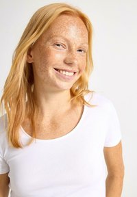 Hvid kortærmet t-shirt med rund halsudskæring. Modellen har lys blond hår og fregnet hud, og smiler svagt.