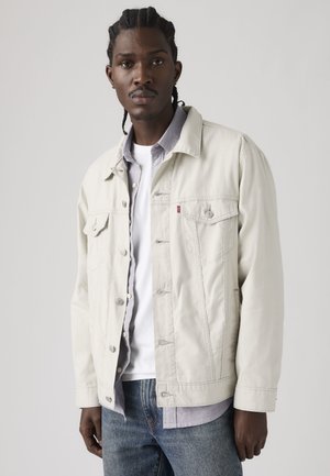 Ung mand med flettet hår iført en lysebeige jakke over en grå skjorte og hvid T-shirt, stående foran en ensfarvet grå baggrund.