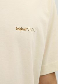 Cream cotton t-shirt featuring a small gold "Originals™ STUDIO" logo on the left side, with a smooth, soft texture and relaxed fit.