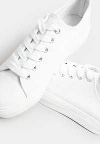 Vita canvas sneakers med platta snören, silverfärgade öglor, gummisulor och en strukturerad finish. Rund tå och minimalistisk design.