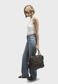 Borsa in pelle marrone con molte tasche con cerniera, dettagli in metallo dorato, portata con due manici. Modella che indossa una canotta chiara e jeans larghi.