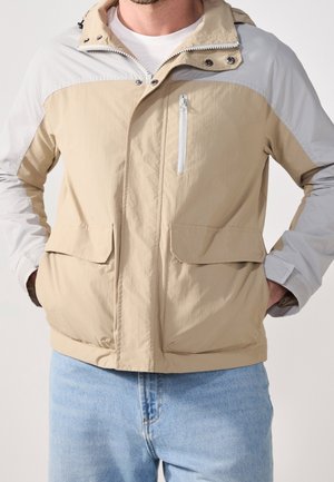 Homme portant une veste à capuche beige et gris clair avec fermeture éclair et boutons, et un jean bleu clair, mains dans les poches de la veste.