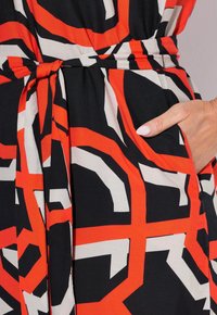 Musta kangasmekko, jossa on abstrakti punainen ja beige kuvio, vyötärölle sidottu ja sivutasku. Luja rakenne ja vartalonmyötäinen siluetti.
