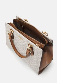 Doekentas in crèmekleurige stof met een geometrisch patroon, accenten van lichtbruin leer, gouden metalen details, dubbele handvatten en een hoofdvak met ritssluiting.