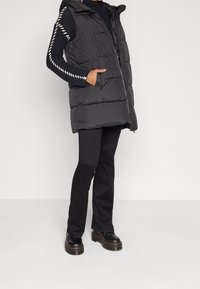 Czarna pikowana kamizelka z kapturem, z poziomym pikowaniem, przednim zamkiem i bocznymi kieszeniami, zestawiona z czarnymi dopasowanymi spodniami i masywnymi butami.