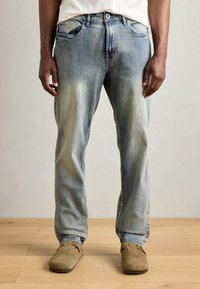 Light blue denim jeans with a relaxed fit, featuring faded accents and subtle distressing. Worn with brown suede slip-on shoes.