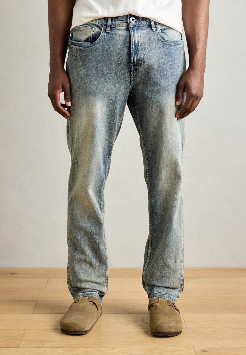 Light blue denim jeans with a relaxed fit, featuring faded accents and subtle distressing. Worn with brown suede slip-on shoes.