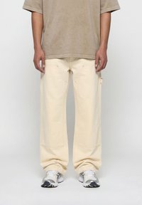 Άντρας που φοράει μπεζ παντελόνι φαρδιάς γραμμής, μπλουζάκι κοντομάνικο ταυπέ και λευκά-γκρι αθλητικά παπούτσια, στέκεται μπροστά σε ένα απλό ανοιχτό φόντο.