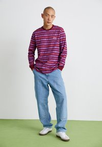 Long-sleeve striped shirt in red and blue with a crew neck; paired with light blue denim pants and white sneakers, set on green flooring.
