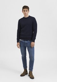 Navy cable-knit sweater with a round neckline, paired with blue jeans and brown Chelsea boots, standing against a plain background.