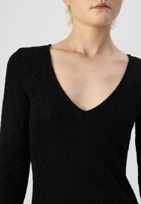 Vestuário preto de manga longa com decote em V, apresentando um tecido texturizado e um padrão de folhas discreto. Acabamento suave e silhueta ajustada.