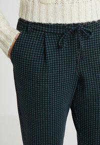Pantalones en un patrón de houndstooth en verde azulado oscuro, con una cinturilla elástica y cordón ajustable. Presenta bolsillos laterales y una tela texturizada.