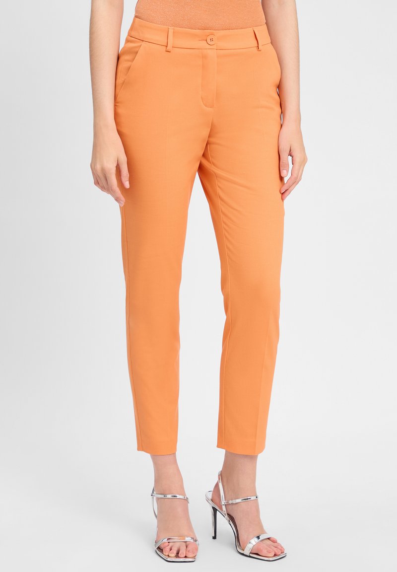 Oranje broek met een getailleerde pasvorm, voorzien van een knoopsluiting, zijzakken en een gladde textuur. Gecombineerd met zilveren sandalen met bandjes.