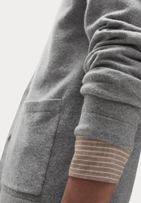Grå sweatshirt med en tekstureret overflade, der har en frontlomme og opkrøllede manchetter, som afslører et stribet mønster i beige og brun.