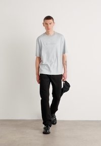 Light gray short-sleeve t-shirt with embroidered logo, paired with black pants and black shoes, standing on a neutral background.