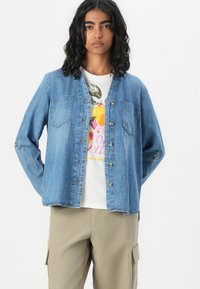 Light blue denim shirt with button closure, two chest pockets, and long sleeves. Worn over a white graphic t-shirt and beige cargo pants.