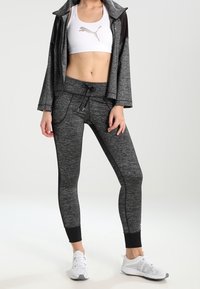 Černý a šedý sportovní outfit skládající se z bundy na zip, přiléhavých legín a sportovní podprsenky s logem. Texturovaná tkanina a zúžený design.