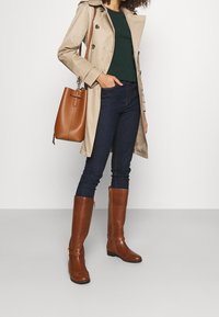 Beige trenchcoat, grön topp, mörka slim fit-jeans och bruna knähöga läderstövlar med guldaccent och sidodragkedja. Brun handväska.