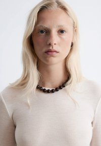 Braune Perlenkette mit glänzenden, runden Perlen. Getragen über einem hellbeigen Hemd mit weicher Textur und klassischem Rundhalsausschnitt.