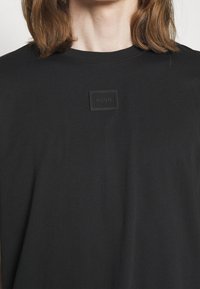 Personne aux cheveux bruns aux épaules portant un t-shirt noir avec un petit patch en relief indiquant "HUGO" sur la poitrine.