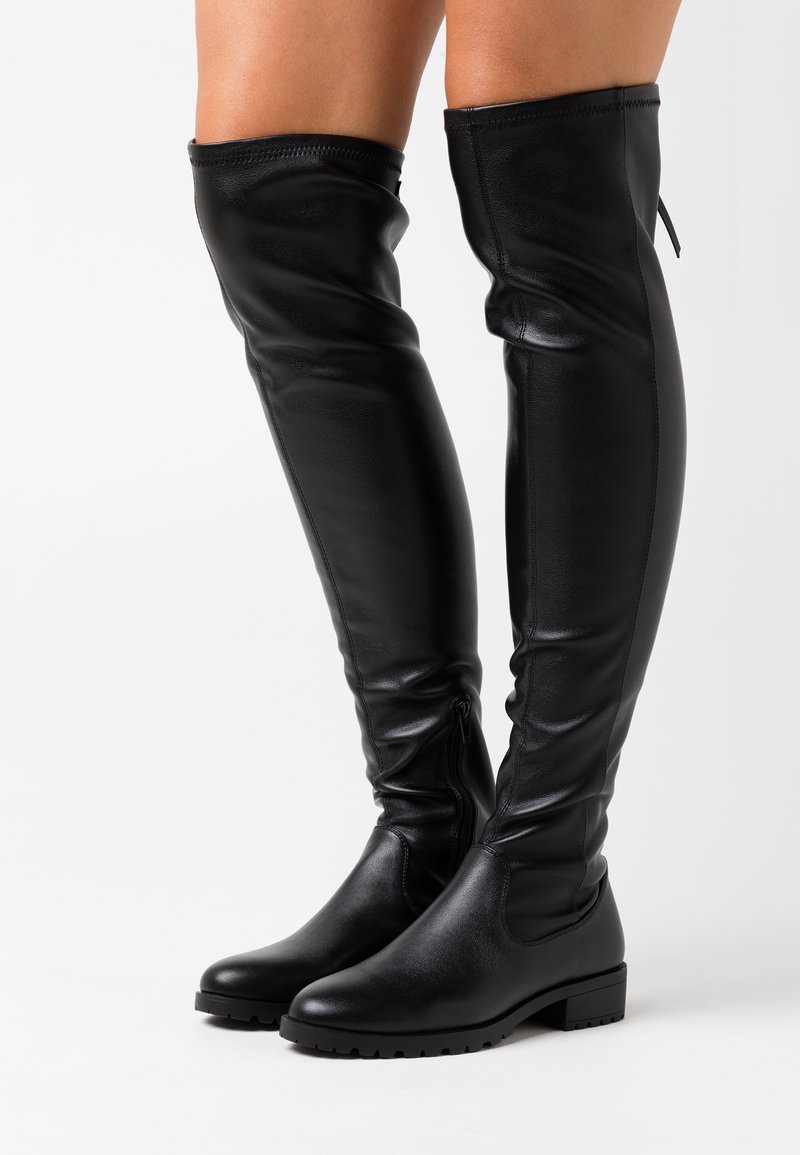 Bottes en cuir noires montant jusqu'aux genoux avec des talons blocs bas et des fermetures éclair latérales, portées sur des jambes nues, présentées sur un fond blanc.