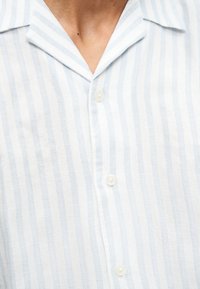 Chemise à col avec rayures verticales bleu clair et blanches, boutonnée sur le devant, portée sur la partie supérieure de la poitrine et le cou.
