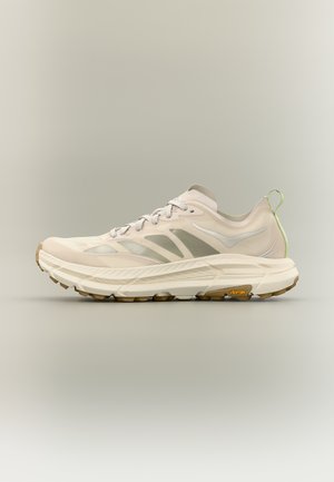 Ανοιχτό μπεζ αθλητικό παπούτσι με λευκές και γκρι λεπτομέρειες, χοντρή μαξιλαρωμένη σόλα, εξωτερική σόλα Vibram και πράσινη υφασμάτινη θηλιά στο πίσω μέρος.