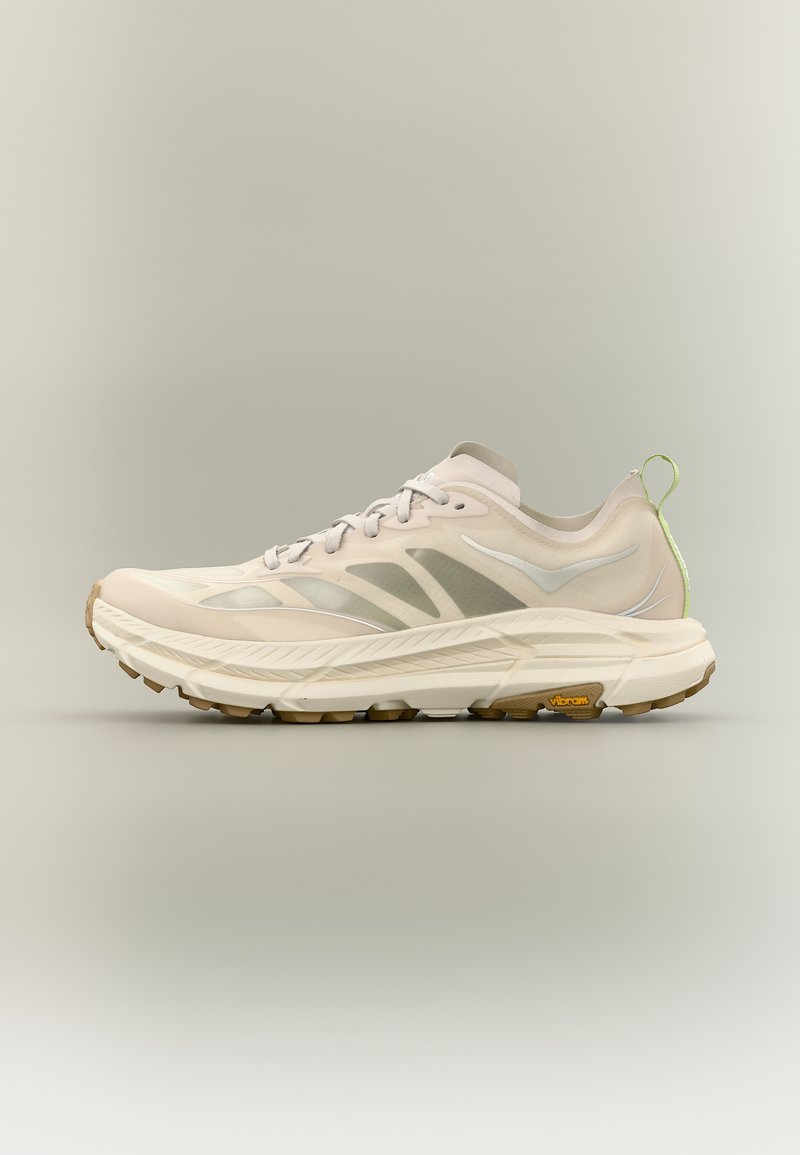 Ανοιχτό μπεζ αθλητικό παπούτσι με λευκές και γκρι λεπτομέρειες, χοντρή μαξιλαρωμένη σόλα, εξωτερική σόλα Vibram και πράσινη υφασμάτινη θηλιά στο πίσω μέρος.