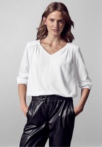 Blusa bianca a scollo a V con dettagli arricciati sulle spalle, maniche lunghe e vestibilità comoda; abbinata a pantaloni neri simili alla pelle con cordoncino.