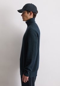 Granatowy sweter z zamkiem przy szyi, o fakturze prążkowanej, z długimi rękawami i luźnym krojem, noszony z dopasowaną ciemną czapką, pokazany z boku.