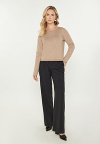 Maglione aderente color marrone chiaro con maniche lunghe, abbinato a pantaloni neri a gamba larga e scarpe nere con punta, presentato su uno sfondo bianco.