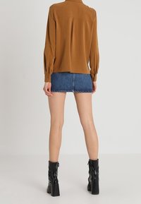 Donna che indossa una blusa lunga marrone e una mini gonna di jeans blu con stivali neri con tacco, in piedi contro uno sfondo neutro.