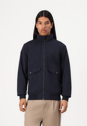 Navy blue jacket with a high collar, front zipper, and two large buttoned chest pockets. Features ribbed cuffs and hem. Made from a lightweight material.