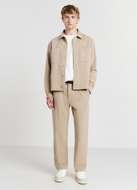 Giacca beige con due tasche frontali, indossata sopra una camicia bianca. Il modello indossa anche pantaloni beige e sneaker bianche. Texture del tessuto liscia.