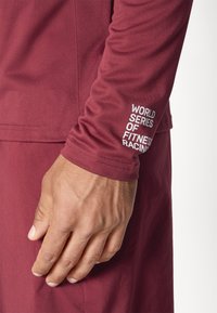 Bordowa, długorękawowa koszulka sportowa z gładką teksturą materiału. Na rękawie znajduje się napis w kolorze białym: "ŚWIATOWE MISTRZOSTWA W WYŚCIGACH FITNESS".