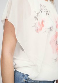 Blusa floreale bianca con motivi rosa e grigi, realizzata in tessuto leggero, con una vestibilità ampia e maniche corte drappeggiate.