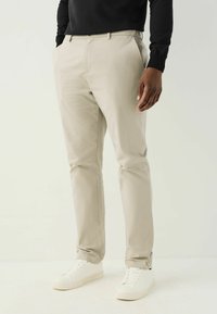 Chino beige in cotone misto, vestibilità aderente con una texture liscia, caratterizzati da tasche laterali e una chiusura con zip, abbinati a sneakers bianche.