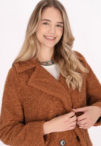 Palton texturat de culoare maro, cu o suprafață pufoasă și blănoasă, prevăzut cu un guler tăiat și nasturi negri, complementat de un top deschis dedesubt.
