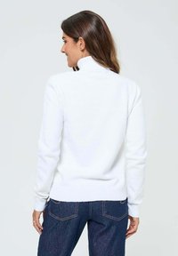 Pull blanc à manches longues avec col haut, en tissu doux. Le dos présente une texture lisse et une coupe ajustée, associé à un jean sombre.