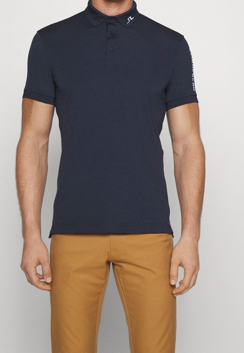 Man som bär en marinblå polo skjorta med logga på kragen och ärmarna, tillsammans med senapsgula byxor, stående mot en enhetlig bakgrund.