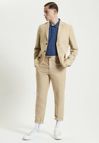 Completo in lino beige con due bottoni, una tasca sul petto, abbinato a un polo blu testurizzato e sneakers bianche. Pantaloni con risvolto e calze visibili.