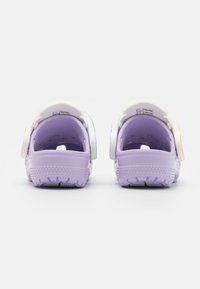 Pareja de zuecos de color lavanda sin talón, con correas ajustables blancas y suelas texturizadas, vistas desde la parte de atrás sobre un fondo blanco.