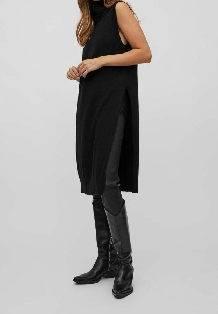 Femme portant une longue tunique noire sans manches avec des fentes latérales, associée à un pantalon noir et des bottes en cuir noires montant jusqu'aux genoux.