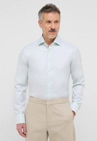 Camicia button-up azzurro chiaro con maniche lunghe e una texture morbida. Presenta un colletto classico e bottoni chiari, abbinata a pantaloni beige.