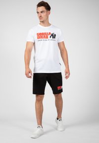 Camiseta de algodón blanca con la impresión gráfica "GORILLA WEAR". Los pantalones cortos negros cuentan con un logo en la pierna derecha. El modelo lleva zapatillas blancas.