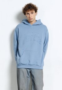 Młody mężczyzna z kręconymi włosami, ubrany w jasnoniebieską bluzę z kapturem z napisem "LOCAL HEROES" oraz jasne spodnie dżinsowe, na białym tle.